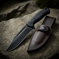 Tactical fixed blade survival knife with black rubber grip and leather sheath on stone surface